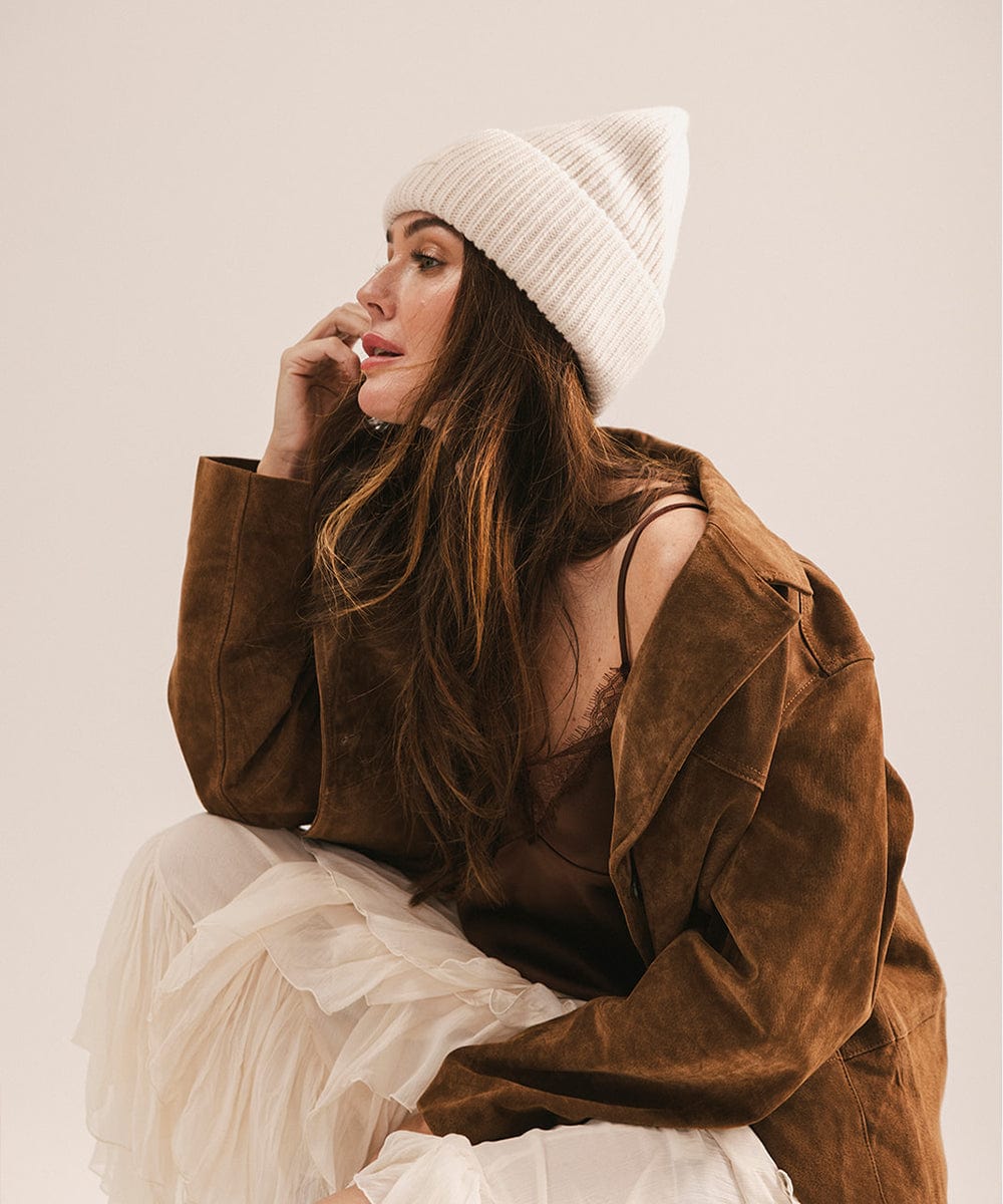 Woman wearing a brown jacket, white skirt and an off white beanie, facing sideways against a white background. #color_off white