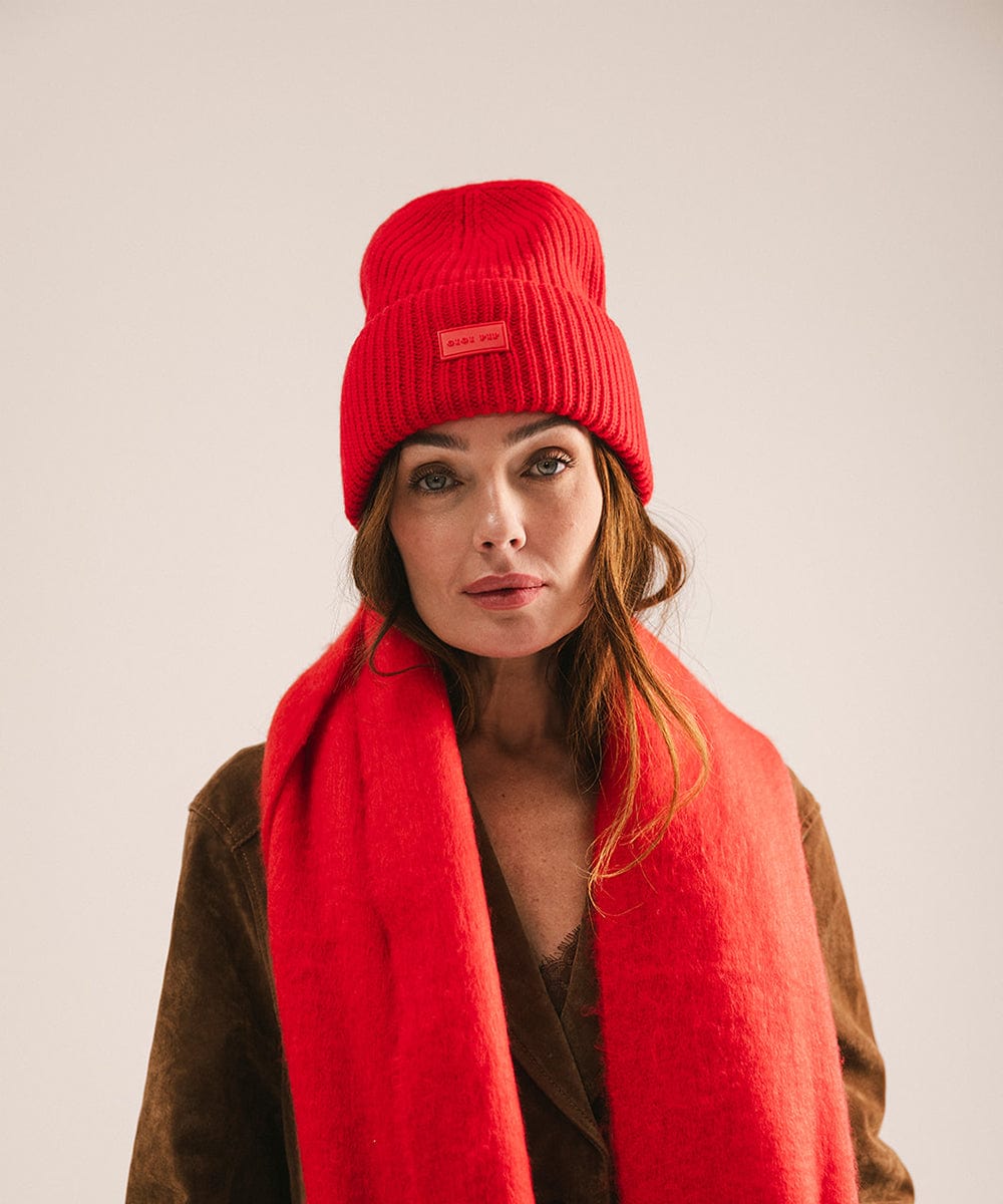 A woman wearing a ruby red colored ribbed beanie and scarf and a jacket against a plain background #color_ruby red