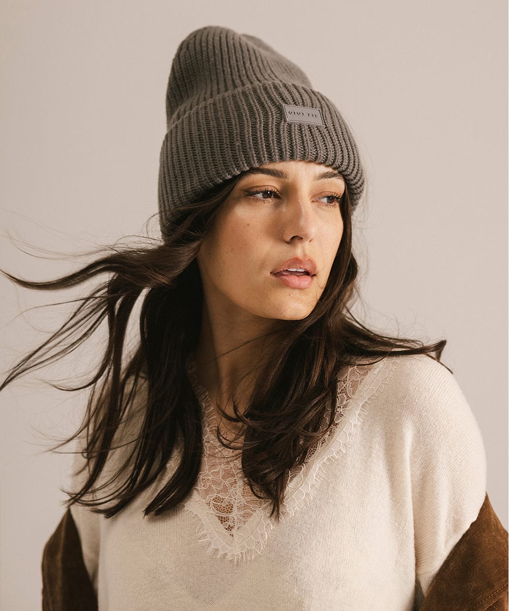 Woman wearing a grey knit beanie with a visible brand logo and a white top, looking sideways against a plain background #color_grey