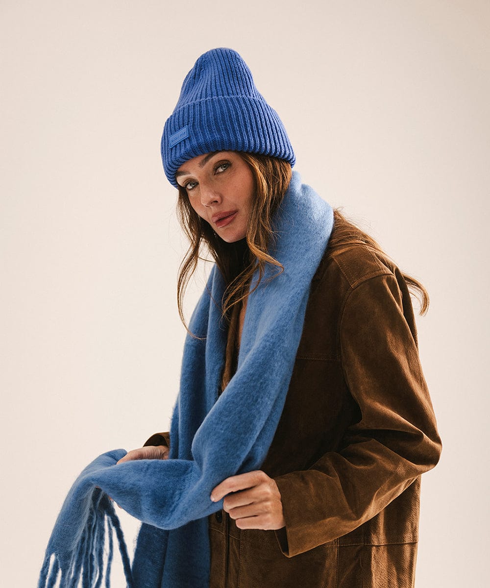 Woman wearing an alpine blue knit hat and scarf and a brown coat against a plain background #color_alpine blue
