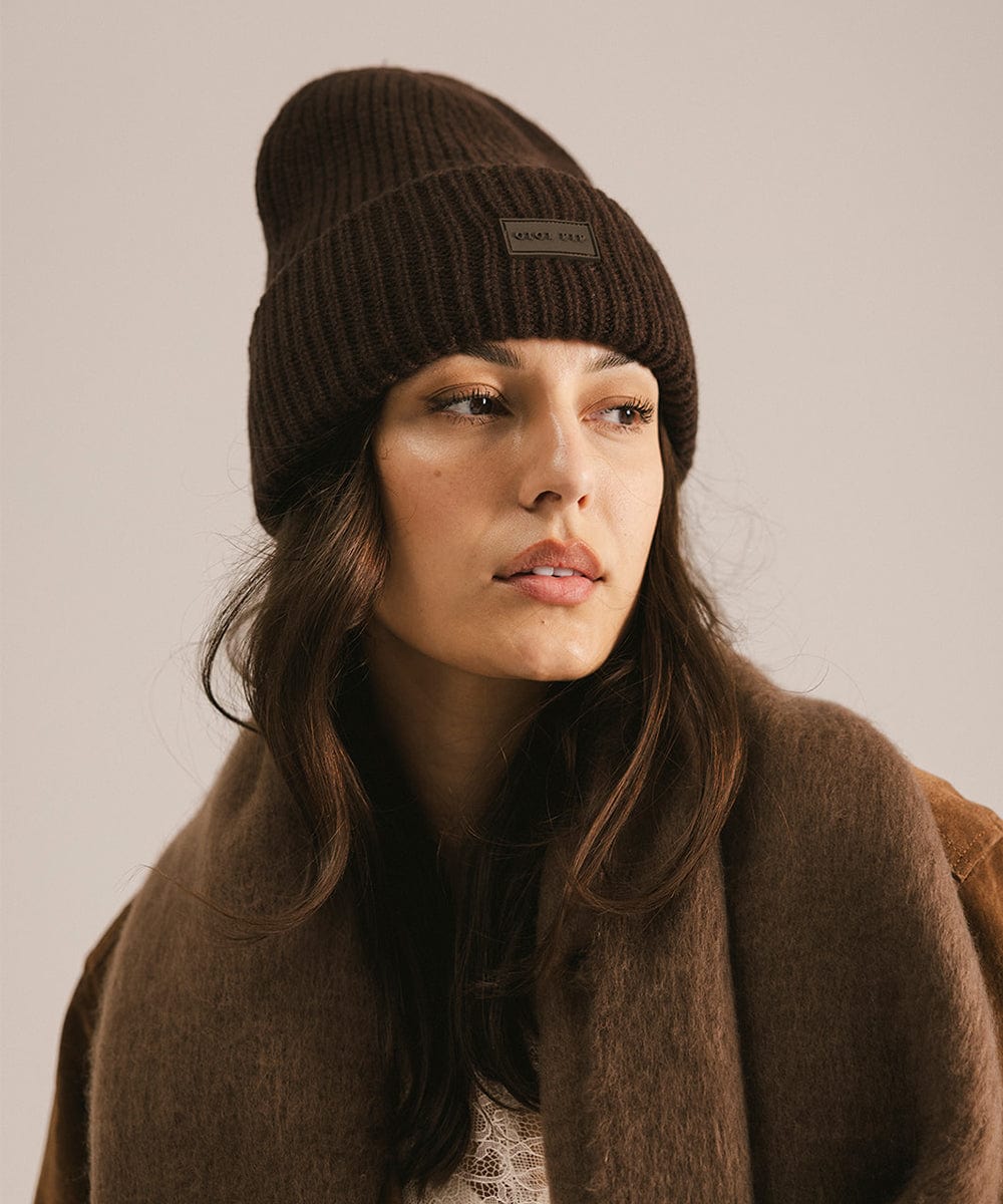 Woman wearing a dark brown knit beanie and a brown coat, looking sideways against a neutral background #color_dark brown
