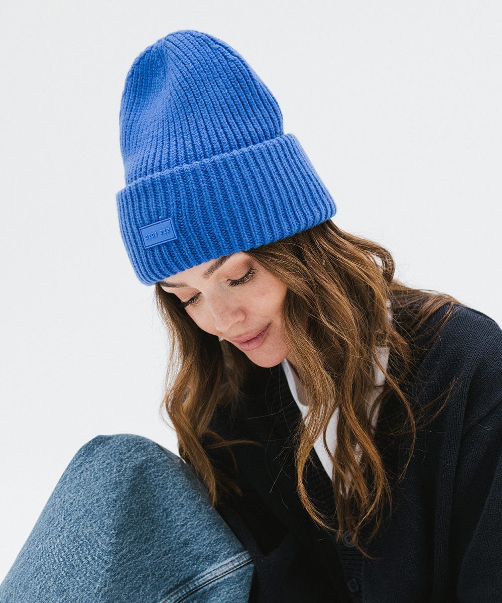 A woman wearing an alpine blue colored ribbed beanie and a dark blue sweater, looking down against a plain background #color_alpine blue