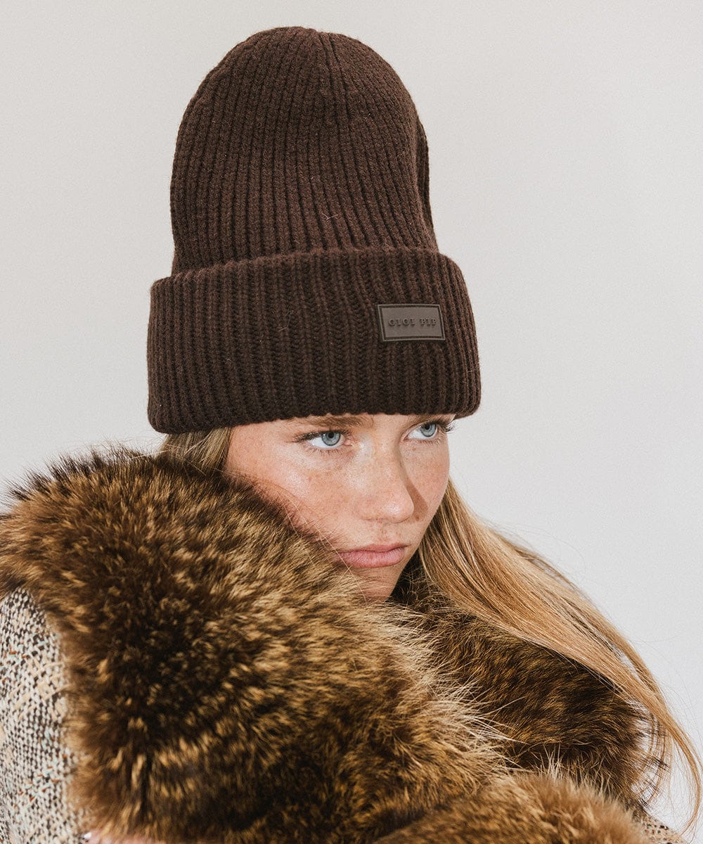 A woman wearing a dark brown chunky ribbed beanie and a brown furry coat, looking sideways against a plain background #color_dark brown