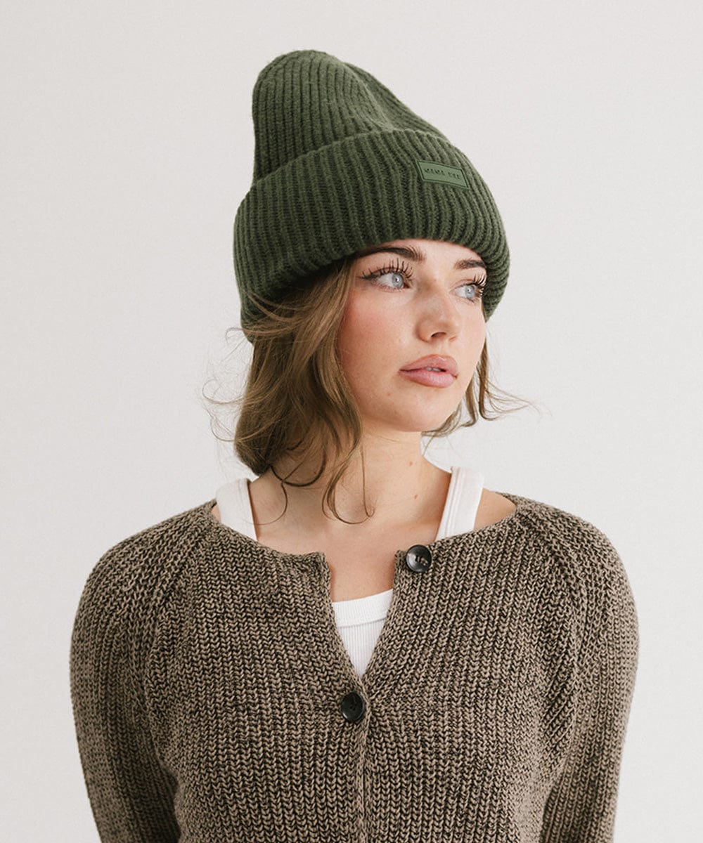 A woman wearing a dark green chunky ribbed beanie and a brown sweater, looking sideways against a plain background #color_dark green