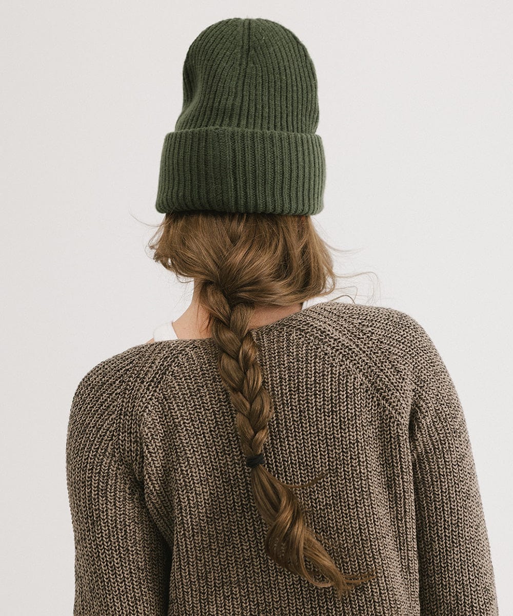A person wearing a dark green chunky ribbed beanie and a brown sweater, facing behind against a plain background #color_dark green