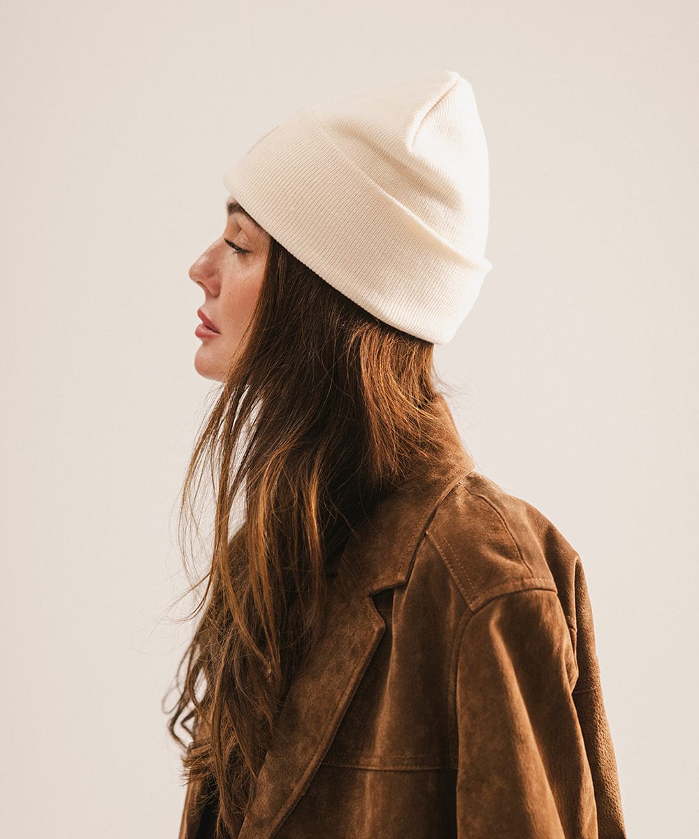 Woman wearing an off white beanie and brown coat, facing sideways against a plain background #color_off white