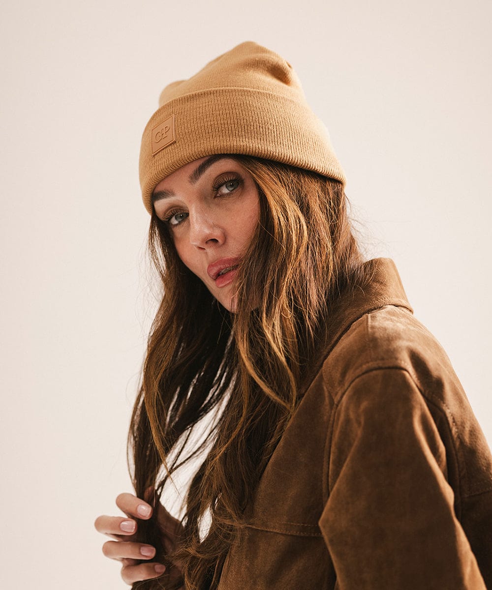 Woman wearing a latte colored beanie and jacket, posing sideways against a plain background #color_latte