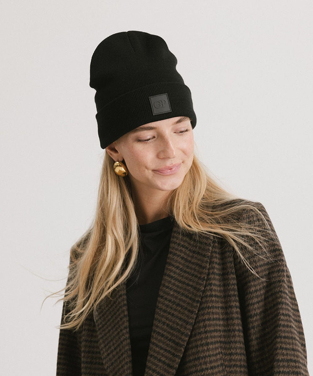 Woman wearing a black beanie with a logo patch and a black t-shirt with a coat, looking sideways against a plain background #color_black