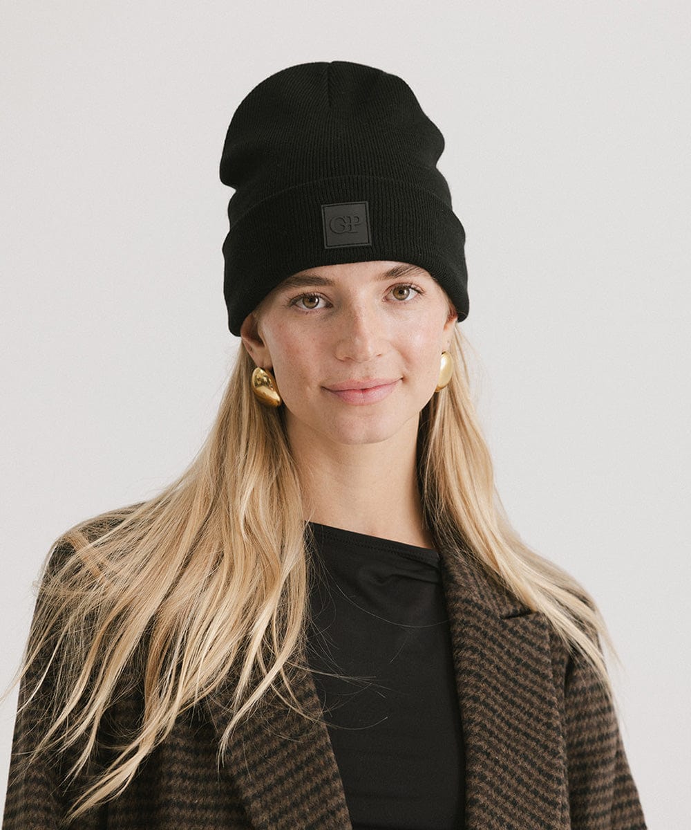 Woman wearing a black beanie with a logo patch and a black t-shirt with a coat against a plain background #color_black
