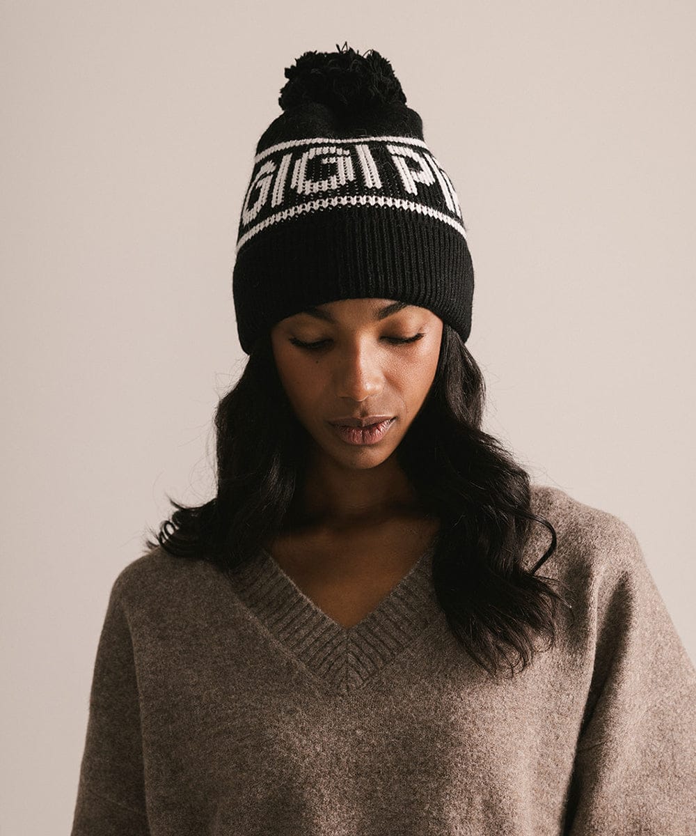 Woman wearing a black beanie with white text and a gray sweater against a beige background #color_black