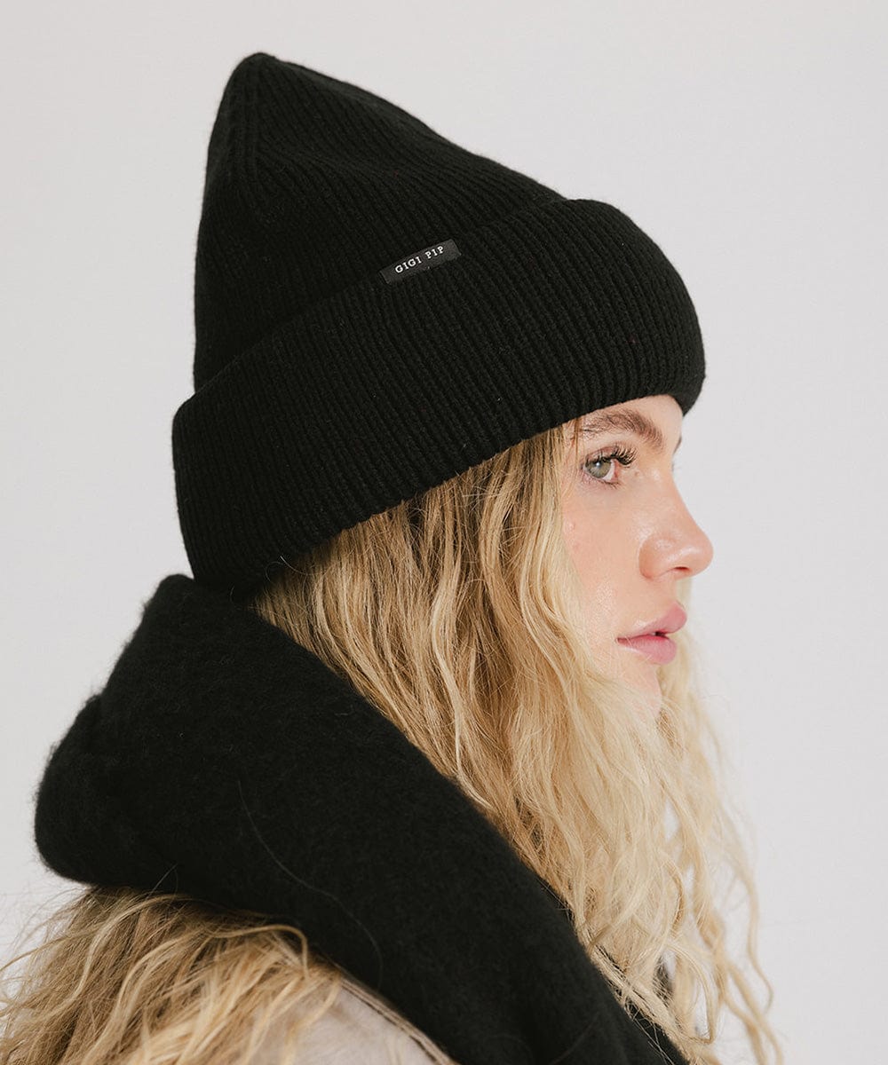 Woman with blonde hair wearing a black oversized knit beanie, facing sideways against a plain background #color_black