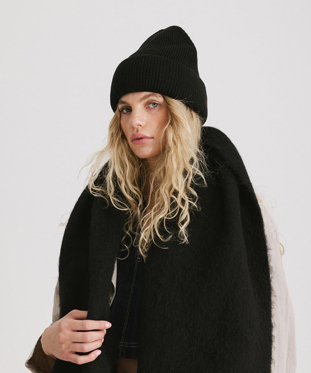 Woman with blonde hair wearing a black oversized knit beanie and a black oversized scarf against a plain background #color_black