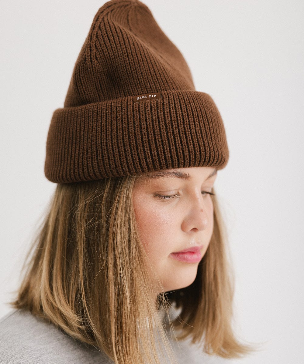 Woman wearing a chocolate colored oversized knit beanie, facing sideways against a plain background #color_chocolate