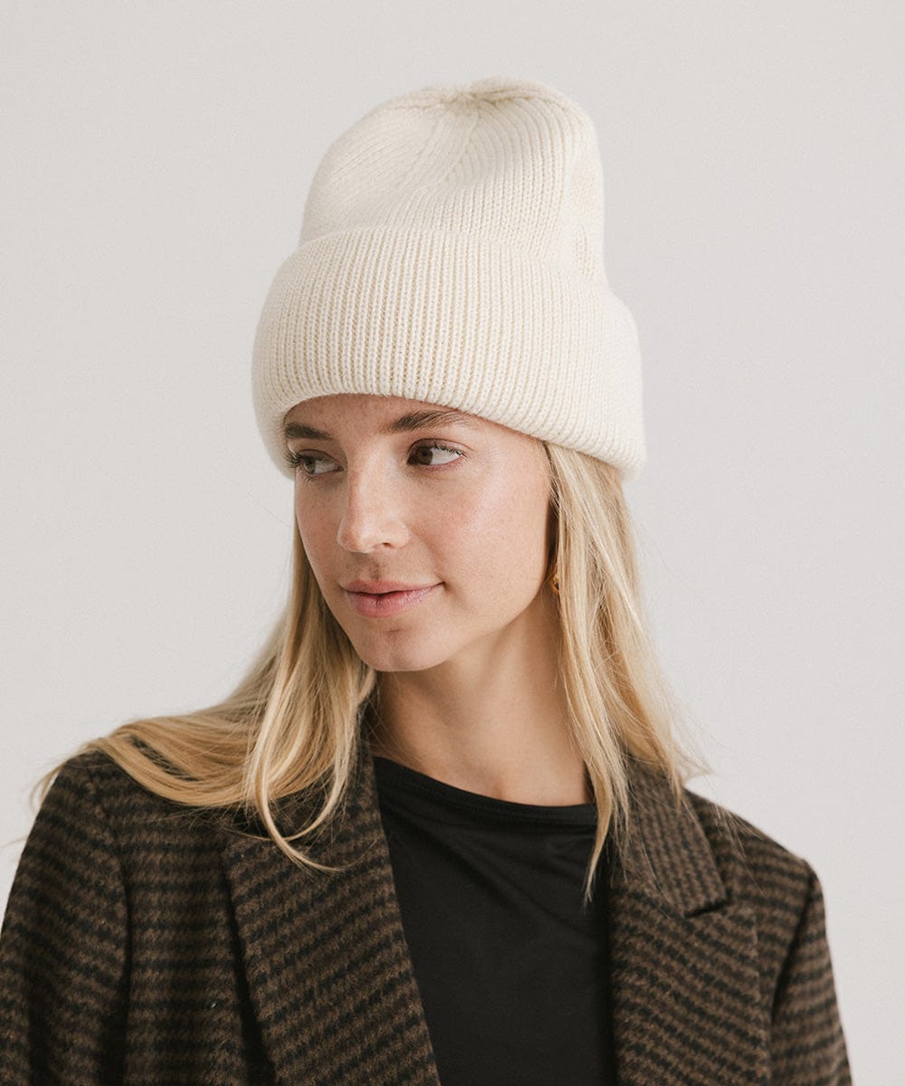 Woman wearing an off white oversized knit beanie and a dark colored coat against a plain background #color_off white