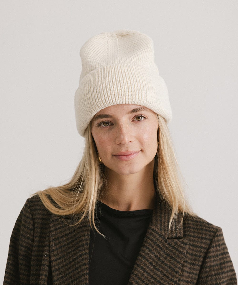 Woman wearing an off white oversized knit beanie and a dark colored coat against a plain background #color_off white