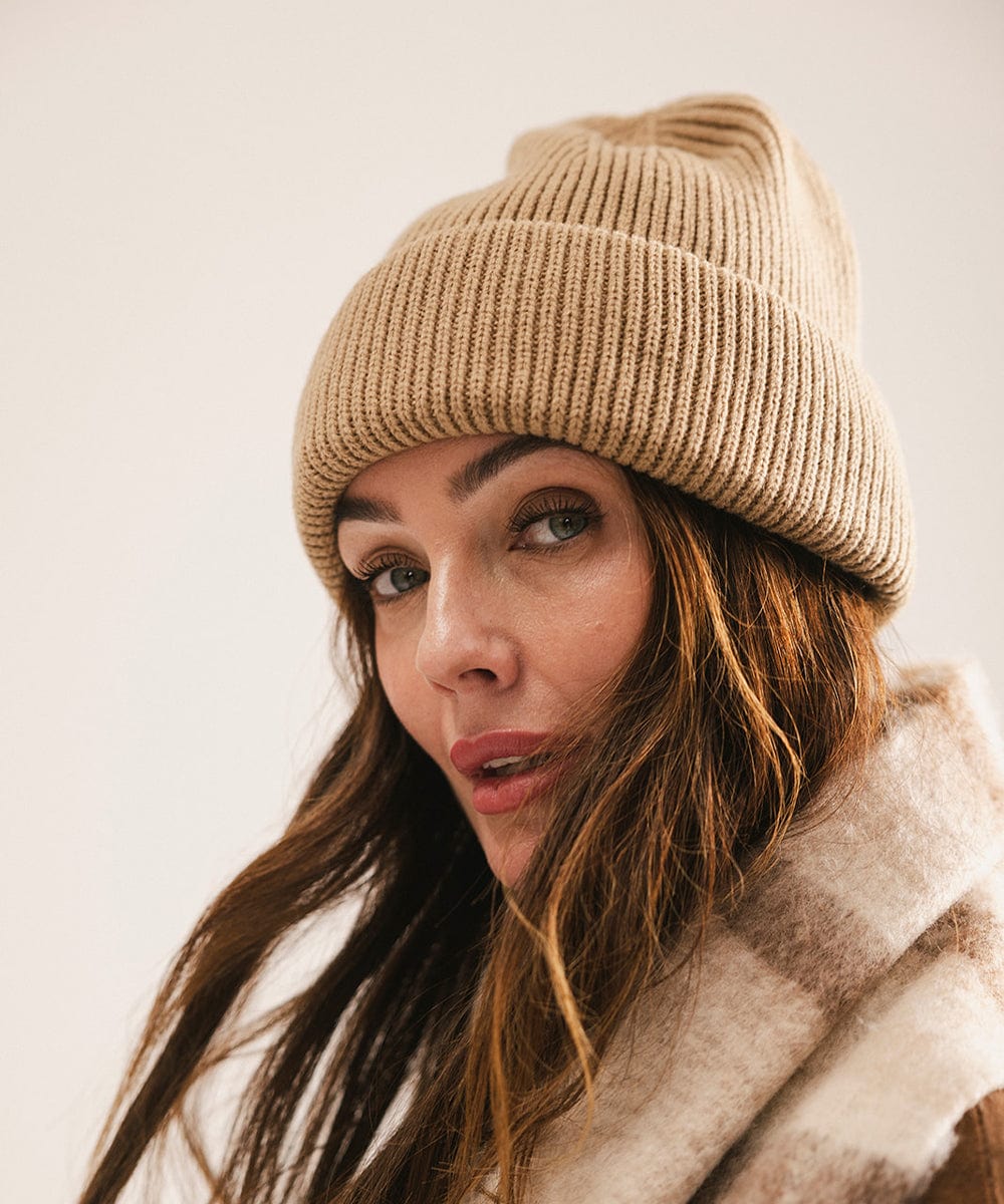 Woman wearing a beige knit beanie and coat against a neutral background #color_tan