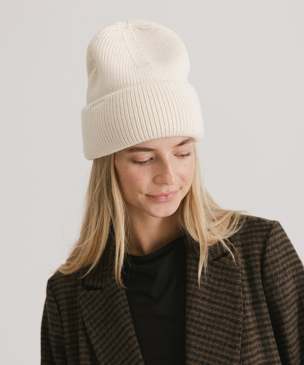 Woman wearing an off white oversized knit beanie and a dark colored coat against a plain background #color_off white