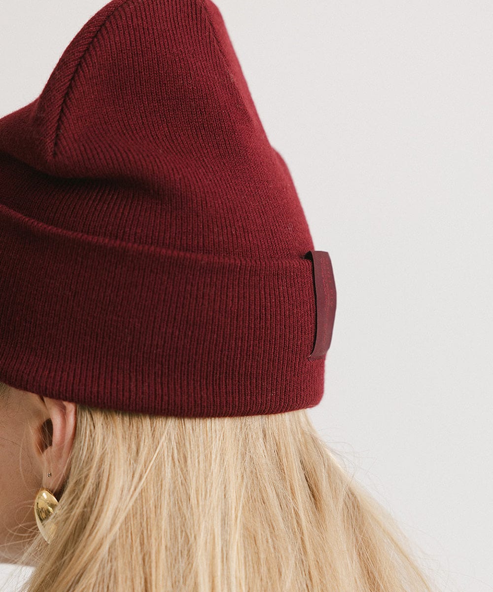 Close up image of a woman with blonde hair wearing a burgundy beanie facing sideways against a plain background #color_burgundy
