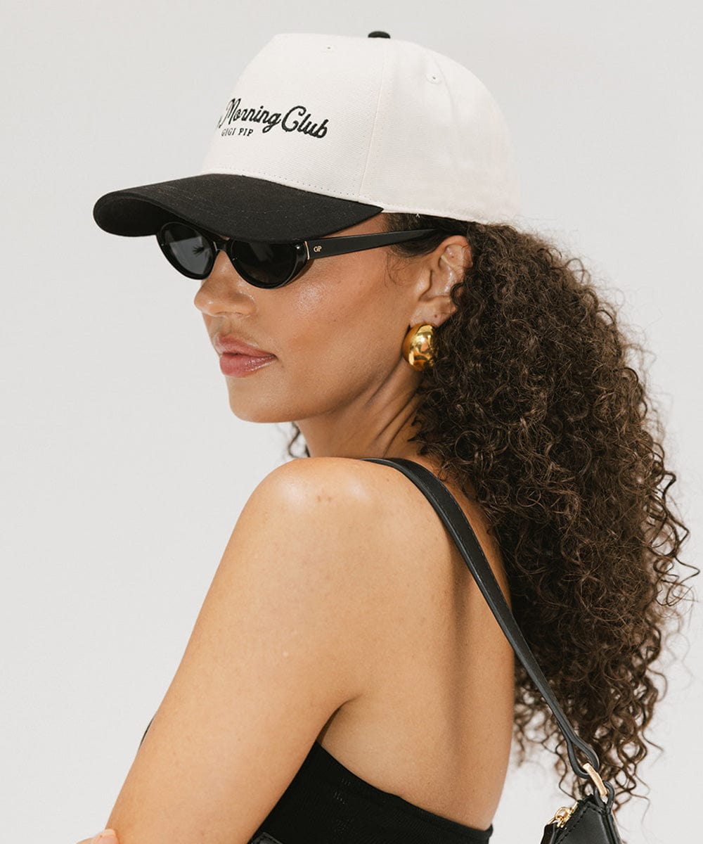 Woman wearing a white cap, black sunglasses and a black top, posing sideways against a plain background #color_black