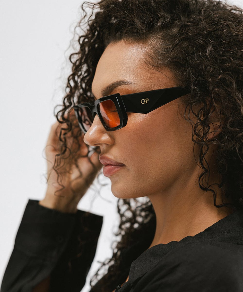 Woman wearing sunglasses with black frame and orange lenses and a black top, facing sideways against a plain background #color_black