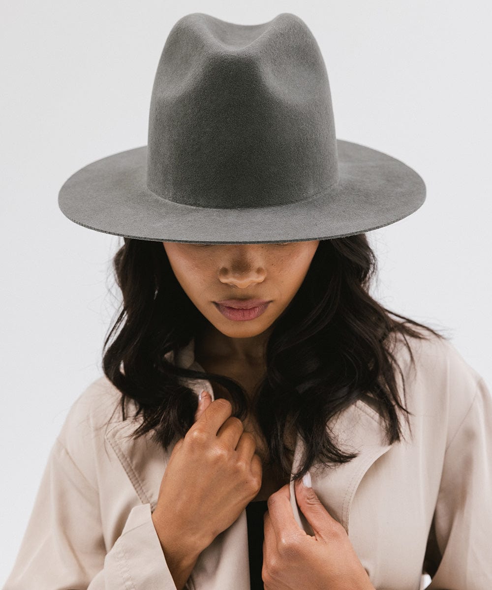Woman wearing a dark grey tall fedora hat and a beige coat against a plain background #color_dark grey