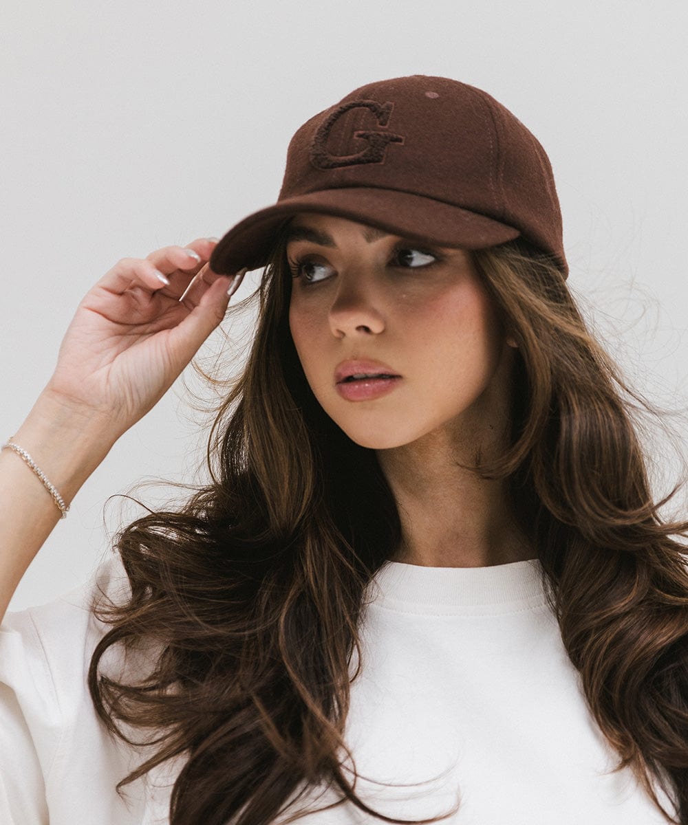 Woman wearing a chocolate brown baseball cap with the letter 'G' and a white top against a plain background #color_chocolate brown