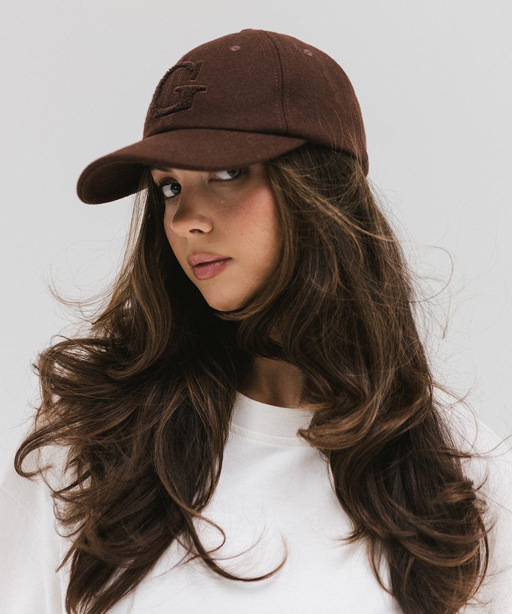 Woman wearing a chocolate brown baseball cap with the letter 'G' and a white top, posing against a plain background #color_chocolate brown