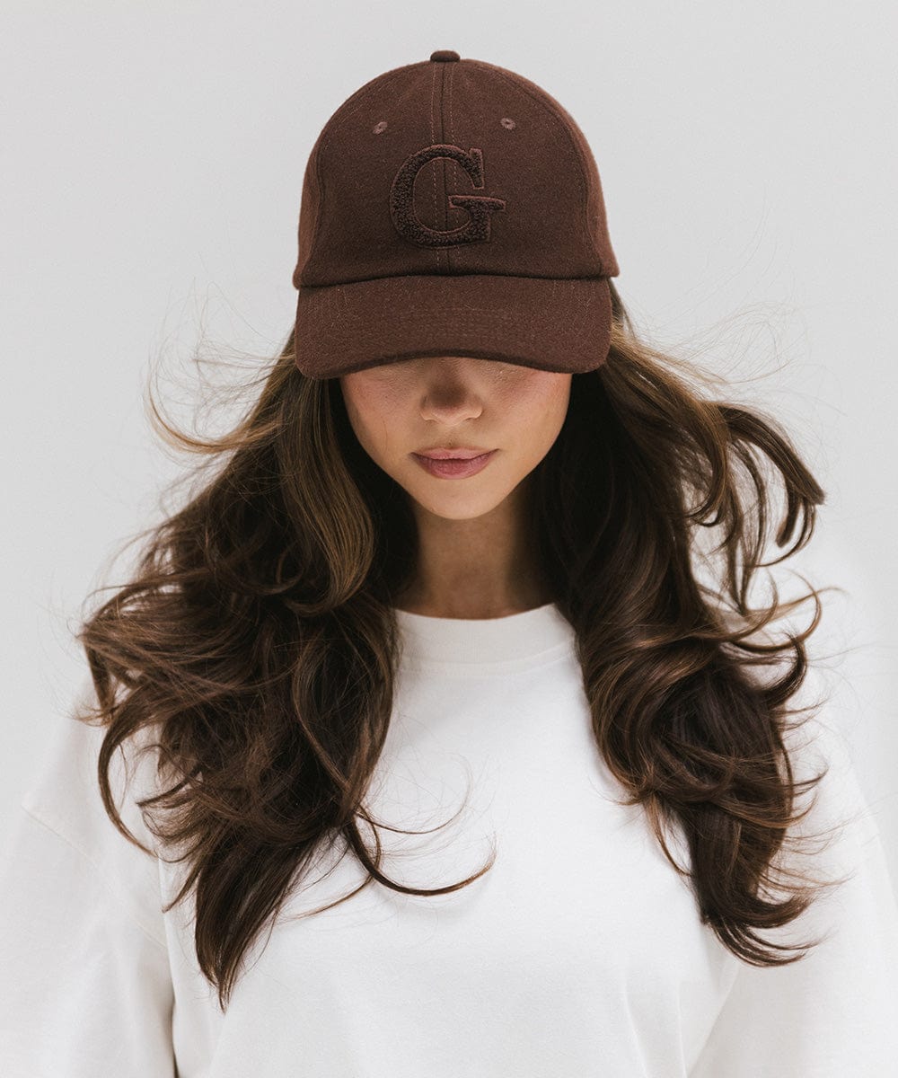 Woman wearing a chocolate brown baseball cap with the letter 'G' and a white top against a plain background #color_chocolate brown