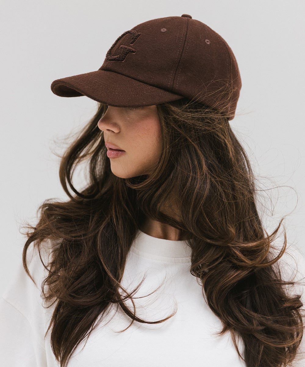 Woman wearing a chocolate brown baseball cap with the letter 'G' and a white top, facing sideways against a plain background #color_chocolate brown