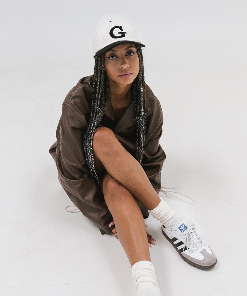 Woman wearing a cream and black baseball cap with the letter 'G', a brown jacket and white shoes, sitting against a plain background #color_cream-black