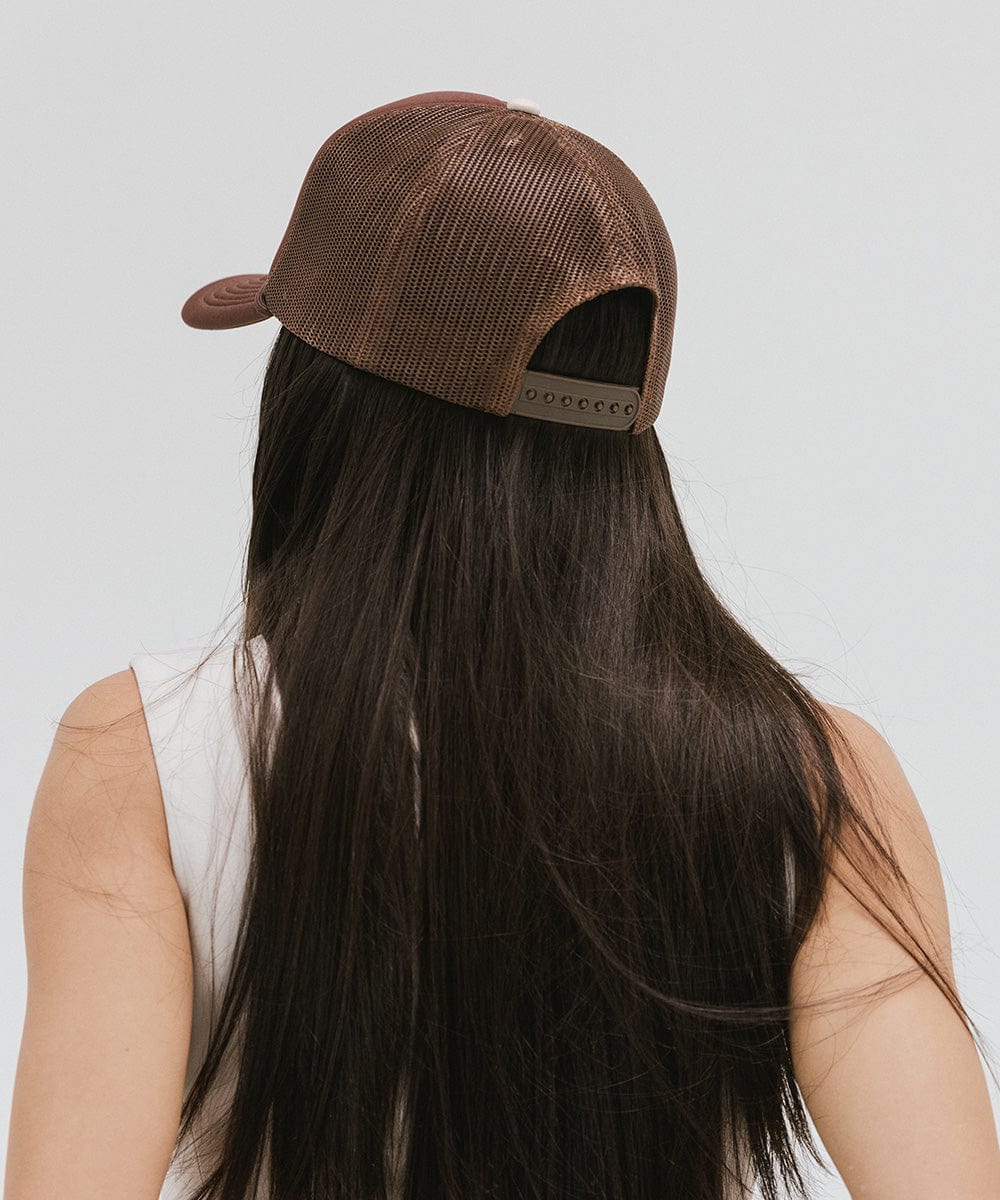 Woman wearing a chocolate brown colored trucker hat facing behind against a plain background #color_chocolate brown