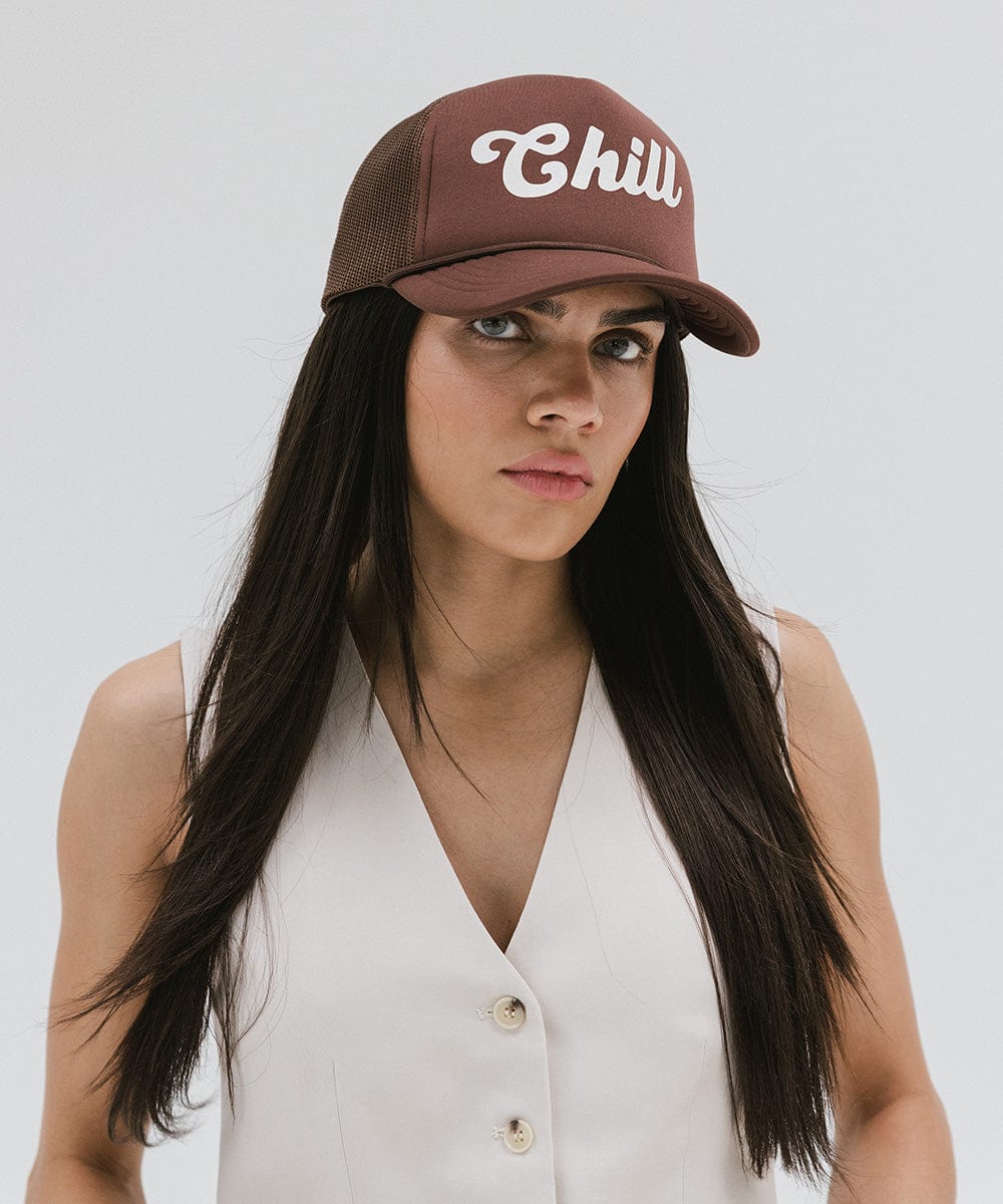 Woman wearing a chocolate brown colored trucker hat with the text 'Chill' and a white top against a plain background #color_chocolate brown