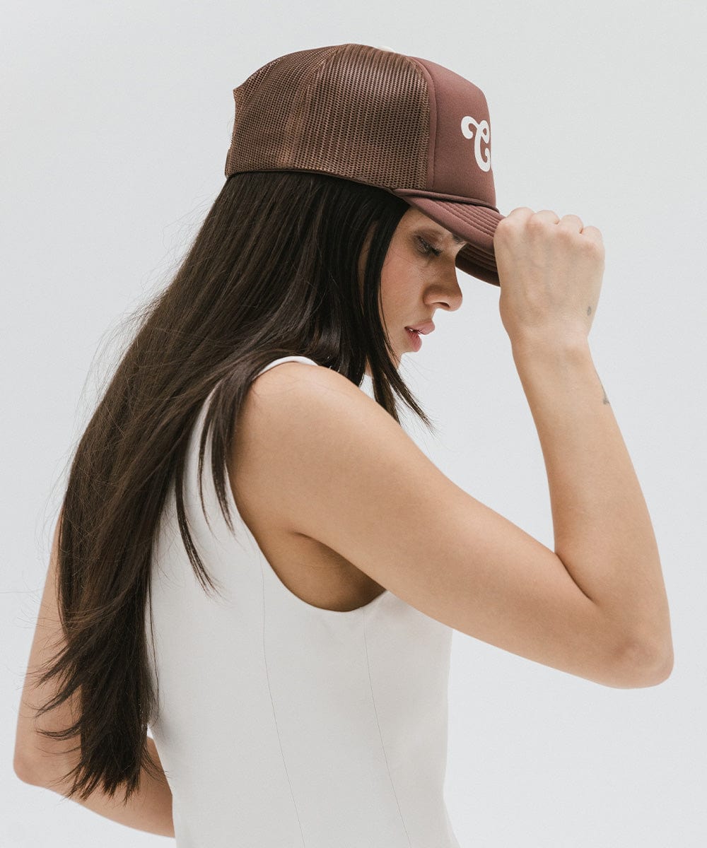 Woman wearing a chocolate brown colored trucker hat and a white top, facing sideways against a plain background #color_chocolate brown