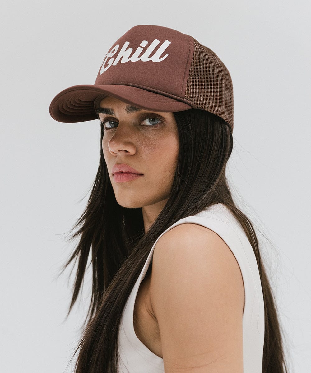 Woman wearing a chocolate brown colored trucker hat with the text 'Chill' and a white top, looking sideways against a plain background #color_chocolate brown