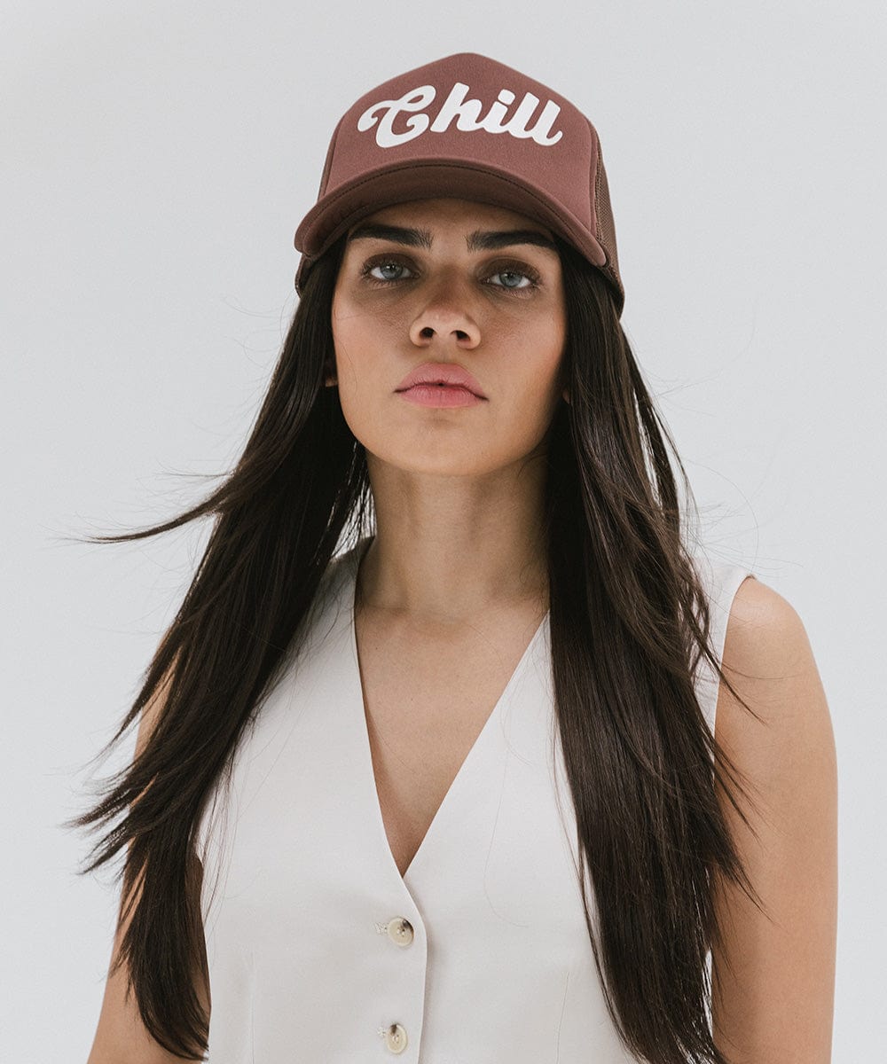 Woman wearing a chocolate brown colored trucker hat with the text 'Chill' and a white top against a plain background #color_chocolate brown