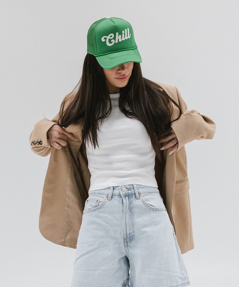 Woman wearing a vintage green colored trucker hat with the text 'Chill', a white t-shirt with a beige blazer and light blue denim shorts against a plain background #color_vintage green