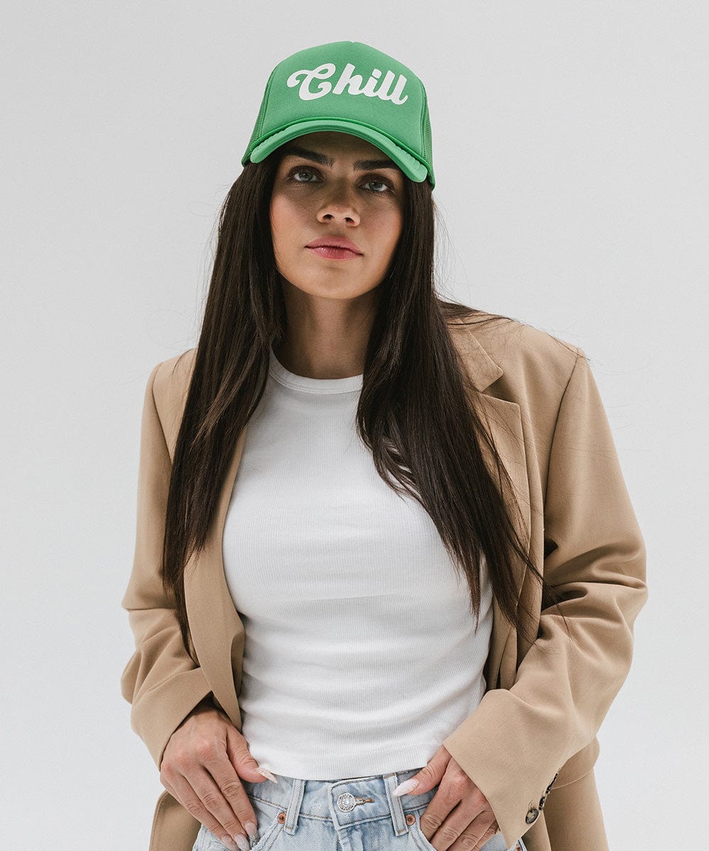 Woman wearing a vintage green colored trucker hat with the text 'Chill' and a white t-shirt with a beige blazer against a plain background #color_vintage green