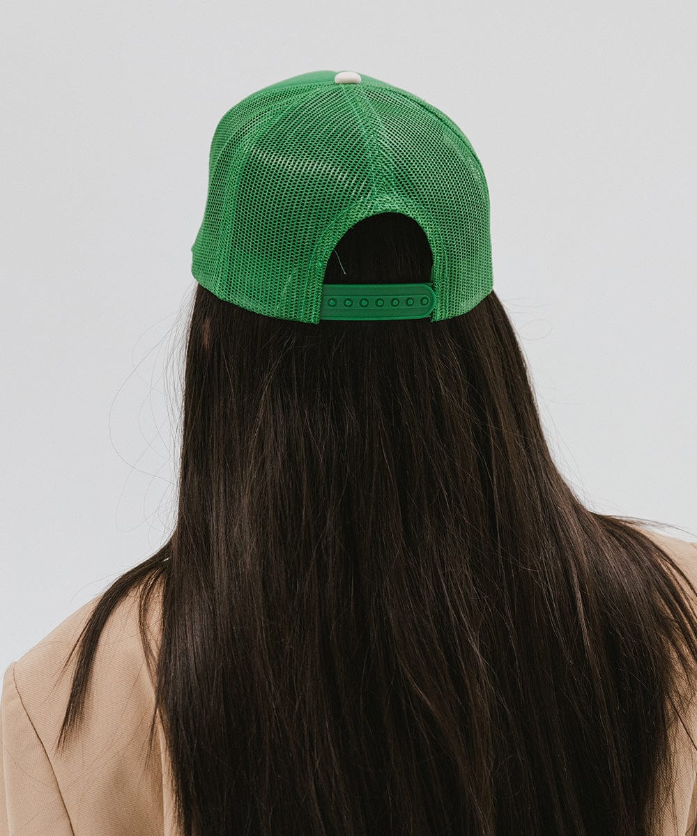 Woman wearing a vintage green colored trucker hat and a beige blazer, facing behind against a plain background #color_vintage green
