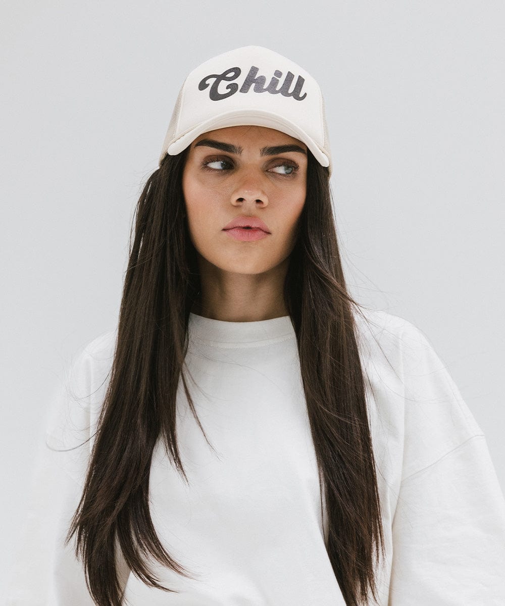 Woman wearing a cream trucker hat with the text 'Chill' and a white t-shirt, looking sideways against a plain background #color_cream