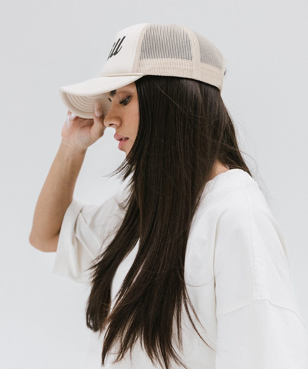 Woman wearing a cream trucker hat and a white t-shirt, facing sideways against a plain background #color_cream