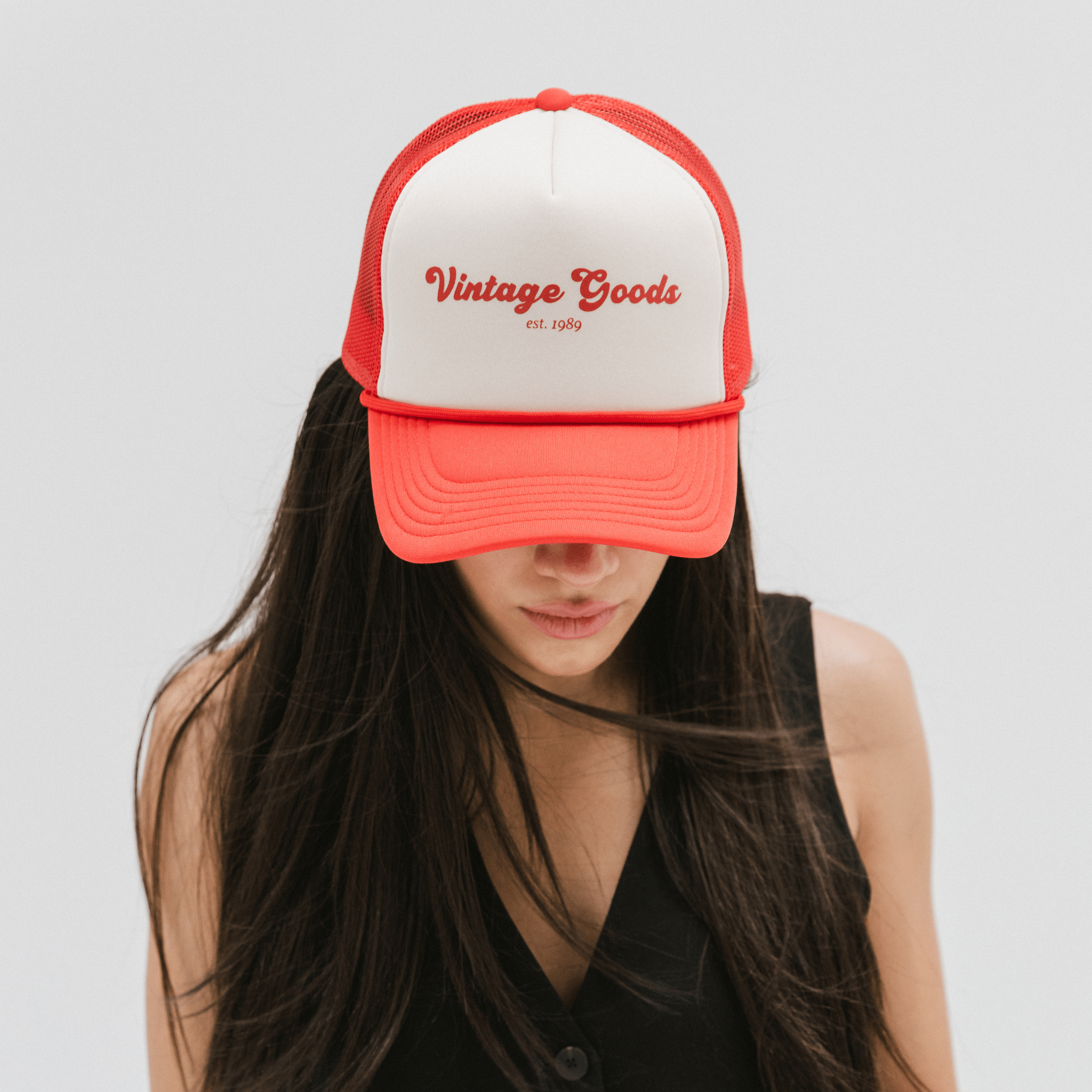 Woman wearing a cream and vintage red colored trucker hat with 'Vintage Goods' text and a black top, looking down against a plain background #color_cream-vintage red