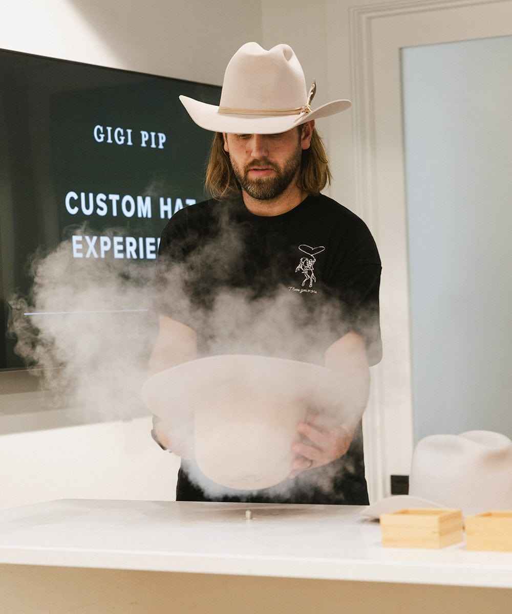 Person wearing a cowboy hat and black shirt with a logo, working with a cloud of smoke in a room with a screen displaying text.