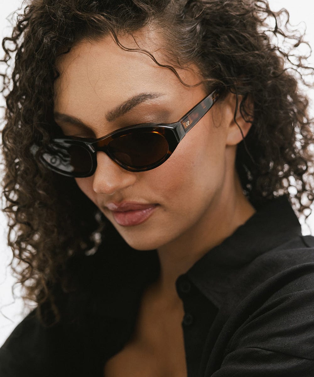 Woman wearing tortoise colored cat eye sunglasses and a black shirt, looking down against a white background #color_tortoise