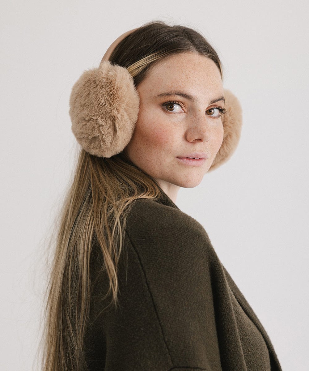 Woman wearing tan fur ear muffs and a brown coat, posing sideways against a plain background #color_tan