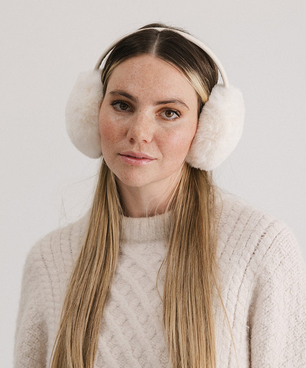 Woman wearing winter white colored fur ear muffs and a white sweater against a plain background #color_winter white