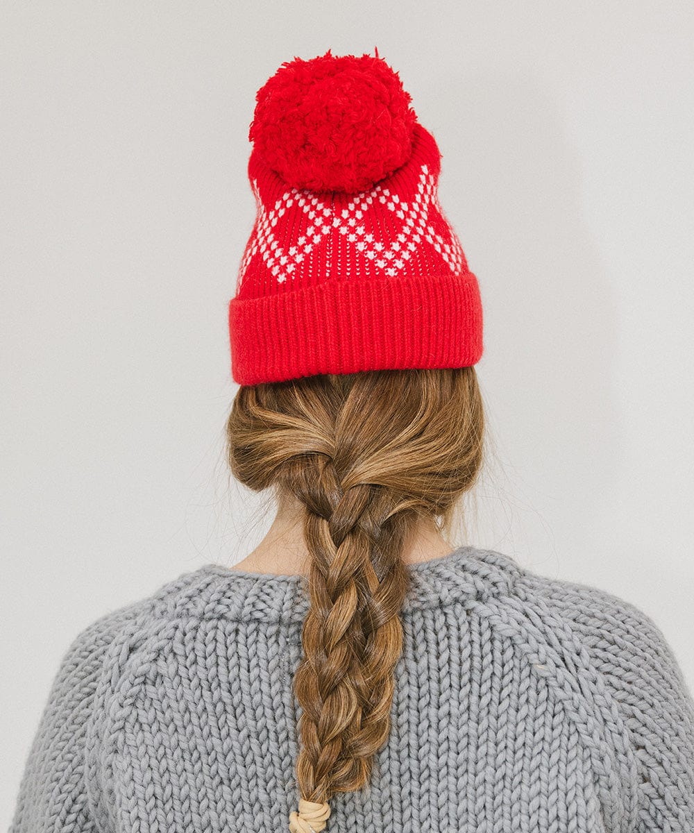 Woman wearing a ruby red colored knit beanie with a pom-pom and a grey sweater, facing behind against a plain background #color_ruby red