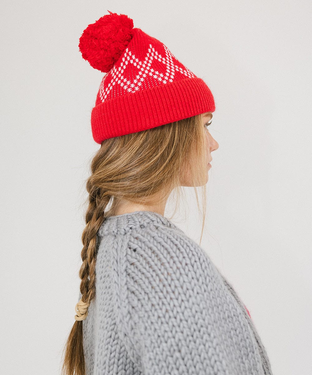 Woman wearing a ruby red colored knit beanie with a pom-pom and a grey sweater, facing sideways against a plain background #color_ruby red