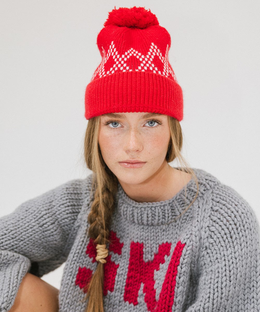 Woman wearing a ruby red colored knit beanie with a pom-pom and a grey sweater against a plain background #color_ruby red