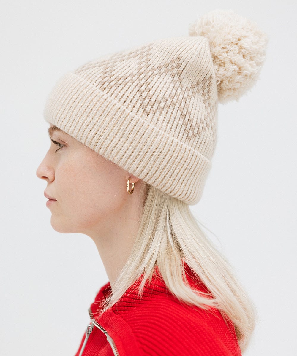 Woman wearing a cream knit beanie with a pom-pom and a red jacket, facing sideways against a plain background #color_cream