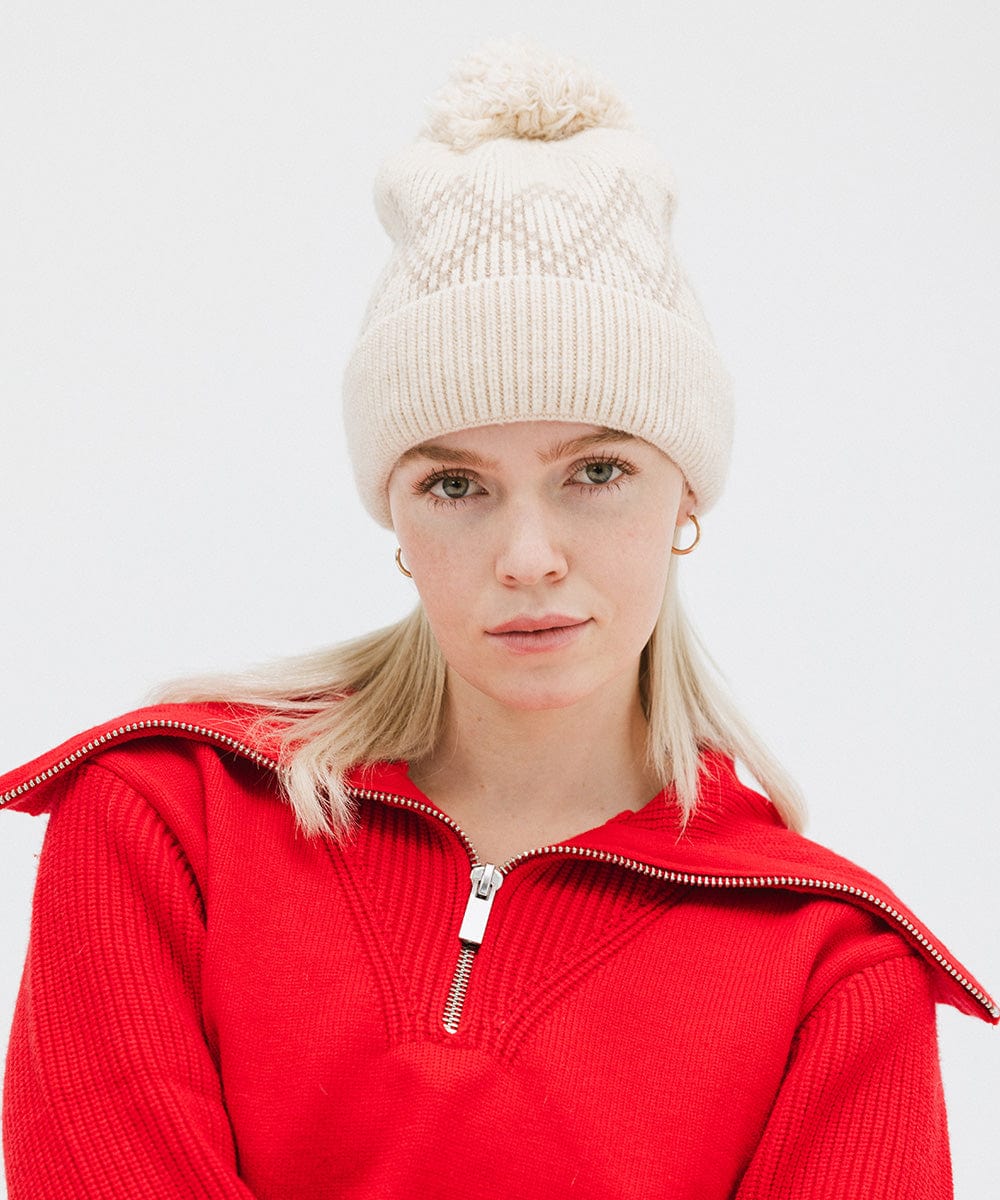 Woman wearing a cream knit beanie with a pom-pom and a red jacket against a plain background #color_cream